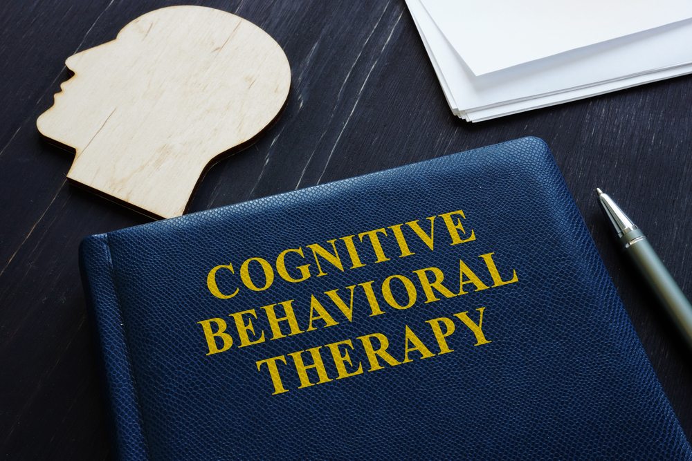 Blue book titled "Cognitive Behavioral Therapy" on a dark wooden desk, accompanied by a wooden head silhouette, papers, and a pen.